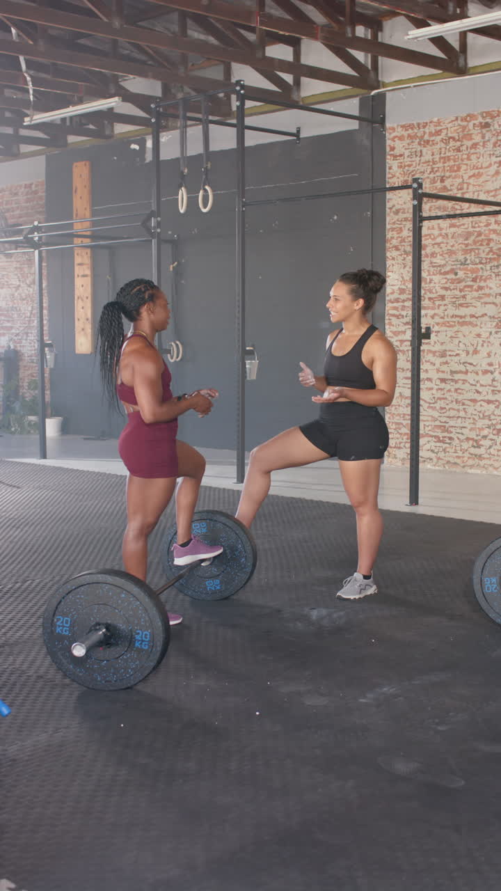 Vertical video: Talking and smiling, two women in gym resting on barbells after workout