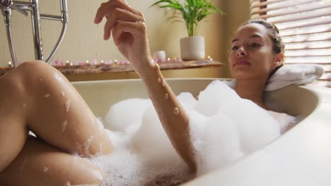 happy biracial woman lying in bathtube with foam and pampering herself with rose petals