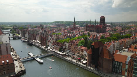 stunning aerial view of gdansk old town district, people crossing the kladka na wyspe spichrzow bridge over nova motlava river - drone pull back, st