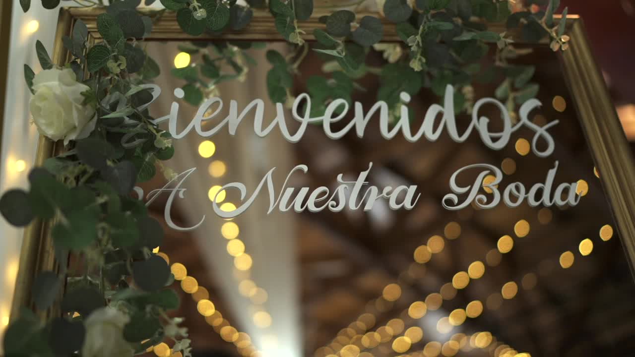 Clip vidéo d’une caméra braquée sur la porte d’un lieu de mariage ornée d’un panneau de bienvenue, de fleurs suspendues et de guirlandes lumineuses en arrière-plan