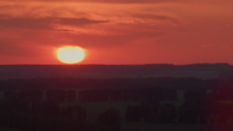 vibrant orange sunset sinking below dark forest horizon, glowing orb illuminating soft clouds with dramatic flare, vast sky awash in warm hues, serene evening silhouette set against open landscape