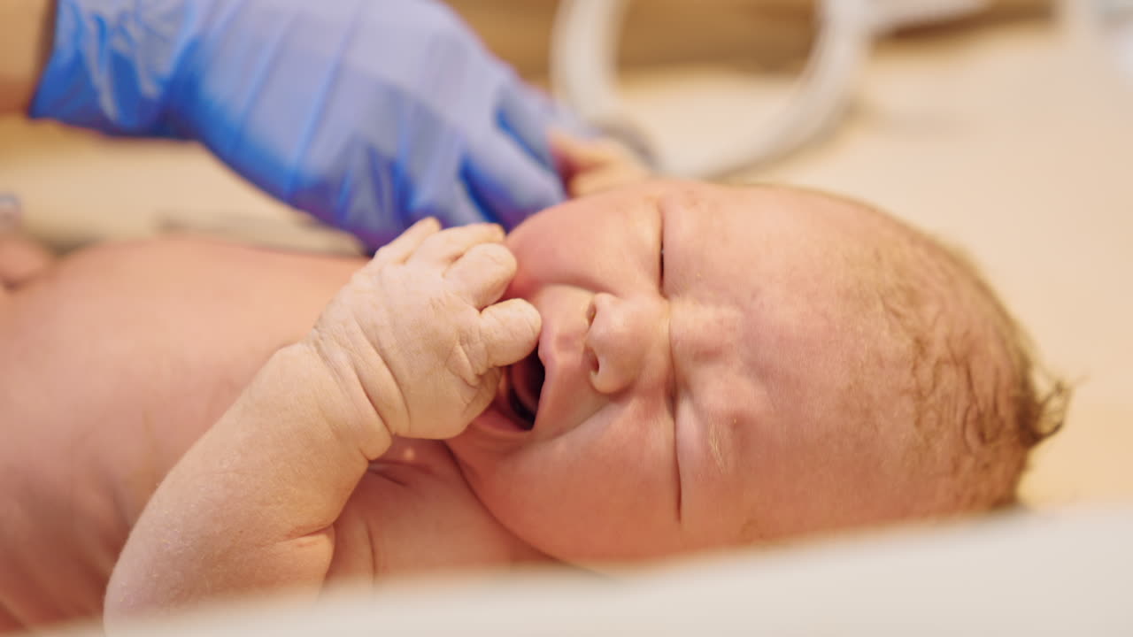 Crying newborn baby on the table. Medic in latex gloves attaches the wire to a child's hand. Close up.