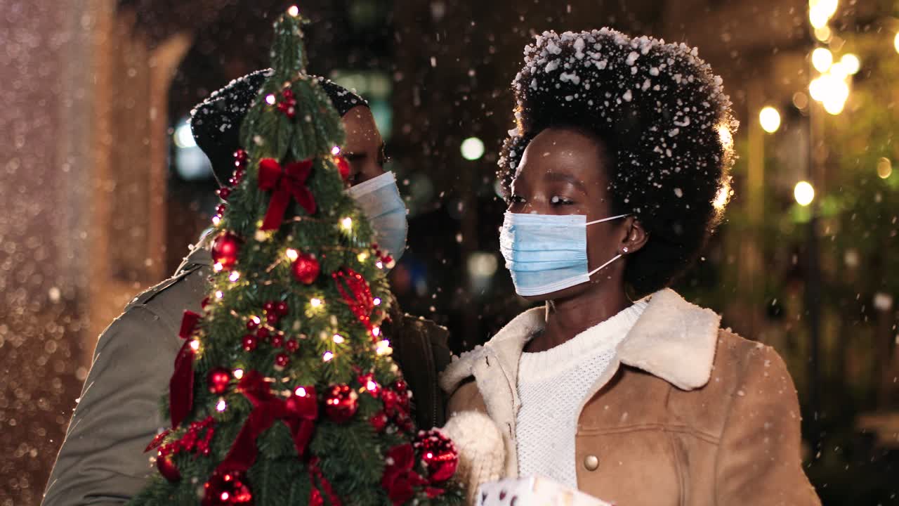 Close-up view of joyful African American couple wearing facial masks talking and holding a Christmas tree while it¬¥s snowing on the street in Christmas