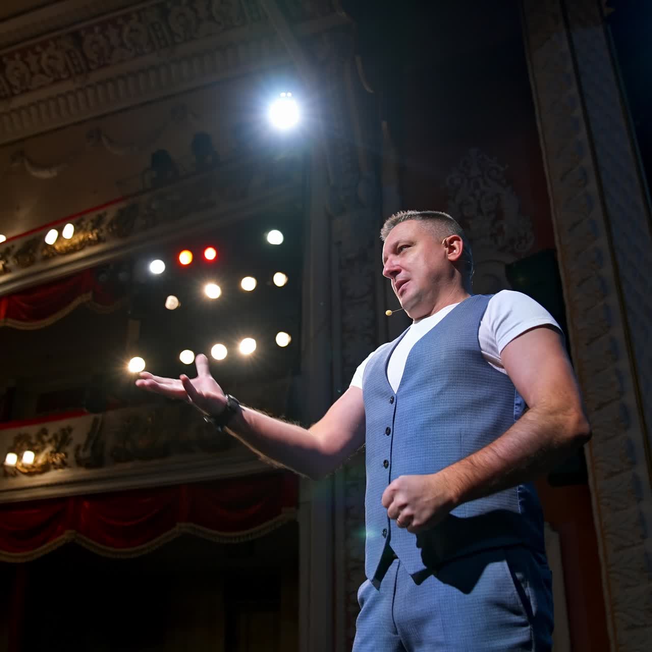 Showman entertains the audience under the light, side view. Big stage. Serious performer speaks from the stage with streched hands