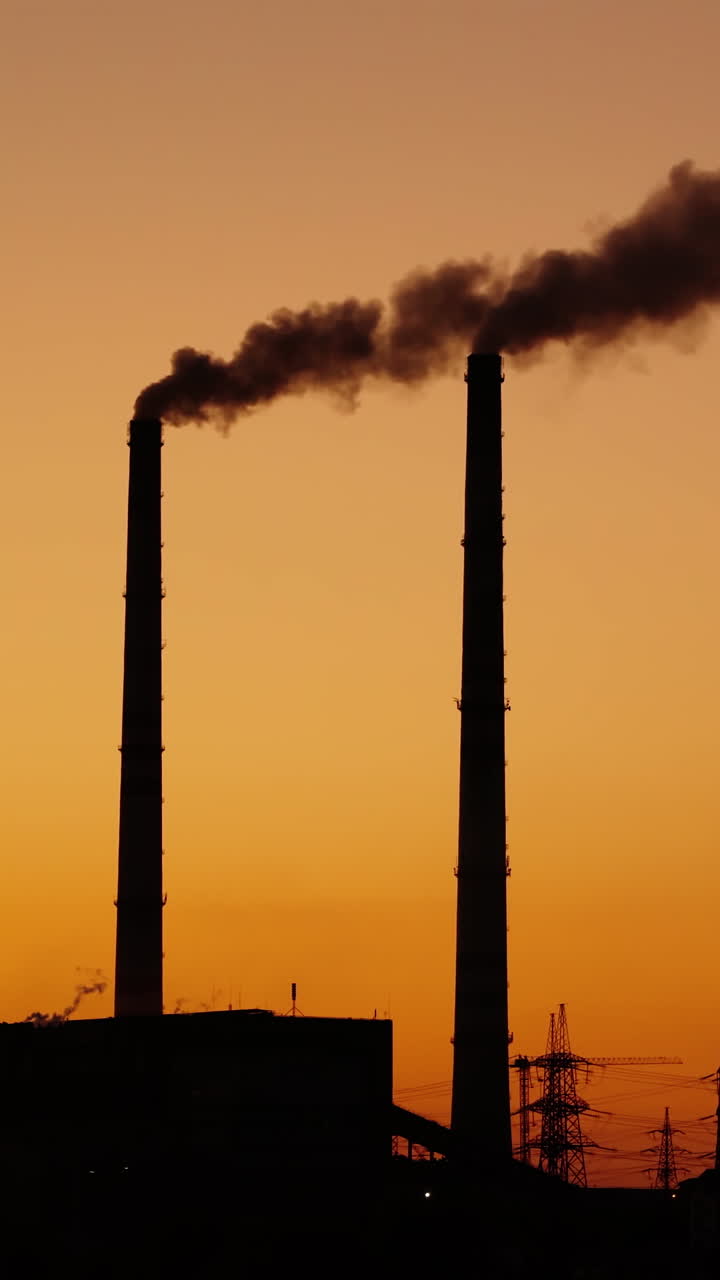 Dark pipes with emissions on the orange sky background. Dirty smoke coming from industrial pipes into the air at sunset. Pollution the environment. Vertical video