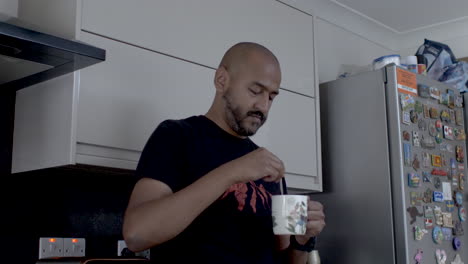 bald adult male indian stirring tea in mug, resting back on kitchen counter