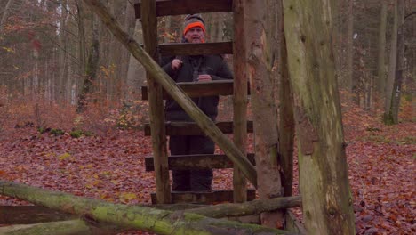 a hunter in a thick winter suit walks up to a high stand, where he observes the surroundings through his binoculars, then climbs up to the high stand