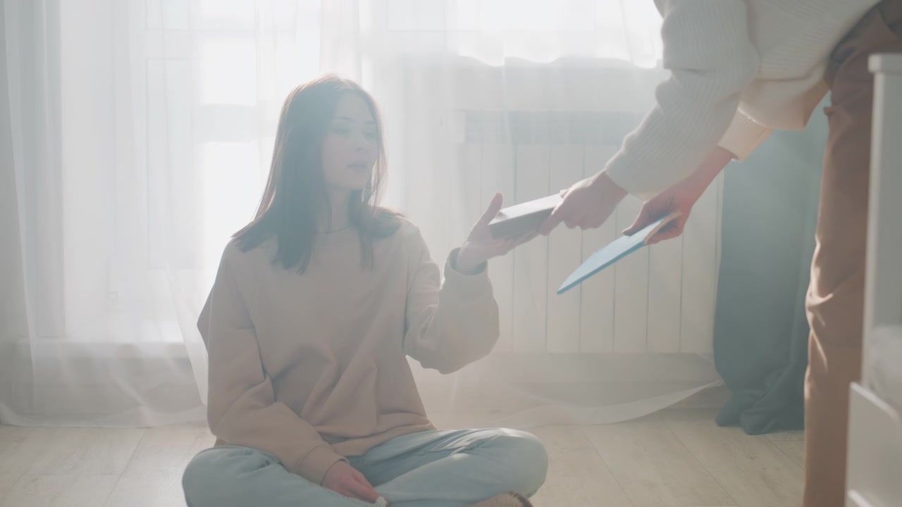 Underviser nærmer sig barfodet og rækker grammatikbog til siddende studerende ved vinduet, intim hjemmecoaching-scene med tæppebelagte trin, hjemmesko, blid vejledning og støttende mentorordning til studiet