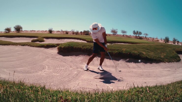 Un golfista colpisce la palla con un ferro da un bunker di sabbia sul fairway in super slow motion, con la sabbia che schizza oltre il bordo erboso sotto un cielo limpido del deserto. La telecamera esegue una panoramica ampia verso destra in 4K a 800fps.