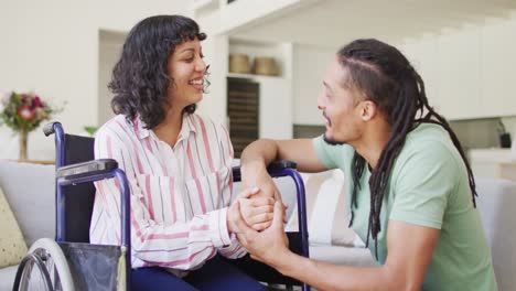 feliz mujer biracial en silla de ruedas y compañero masculino sentado tomados de la mano y hablando en la sala de estar