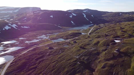 The-top-of-mountain-Vikafjell-with-mountain-road-crossing-and-many-leisure-cabin-huts-up-in-green-hillside---Evening-sunny-aerial-with-beautiful-sunflares