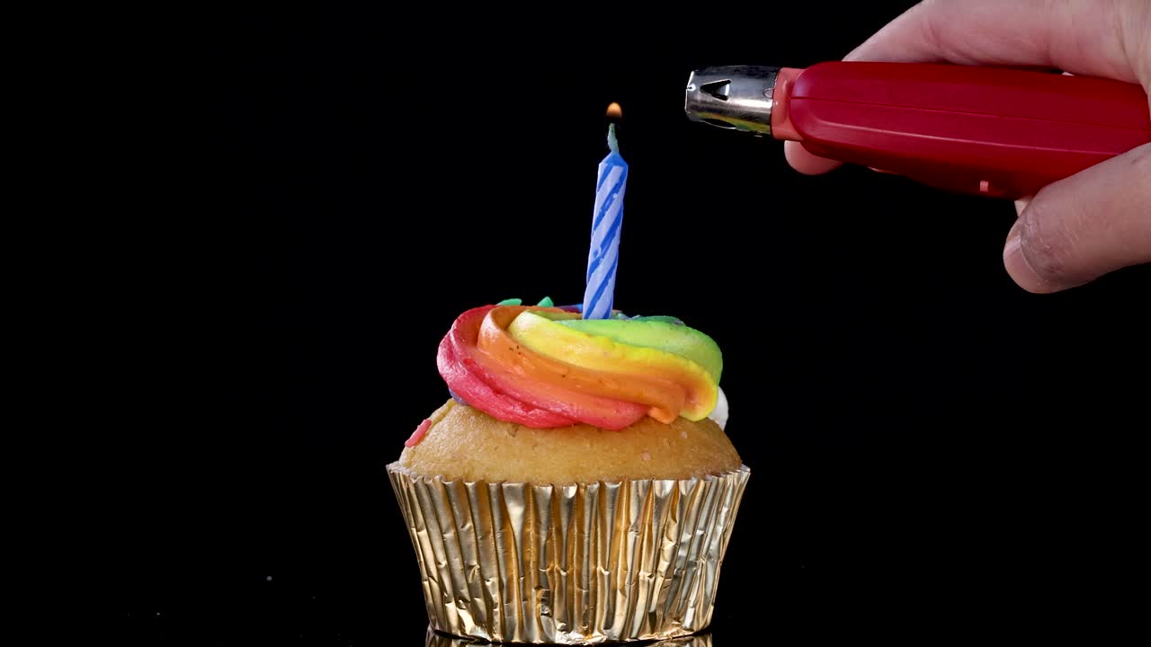 A hand uses a lighter to ignite a blue-striped birthday candle on a rainbow-frosted cupcake, set against a black background with bright, even lighting