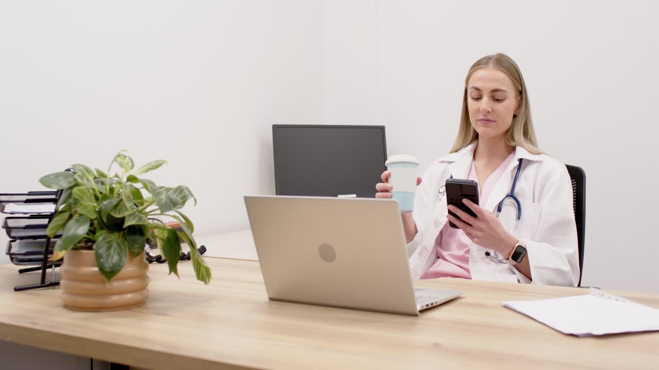 Médica adulta em sua mesa com estetoscópio atendendo o telefone, segurando uma xícara e olhando para o laptop