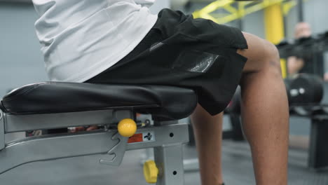 partial view of man wearing white top and black shorts sitting on adjustable gym bench while performing dumbbell curl to build arm muscle inside spacious gym environment with blurred background
