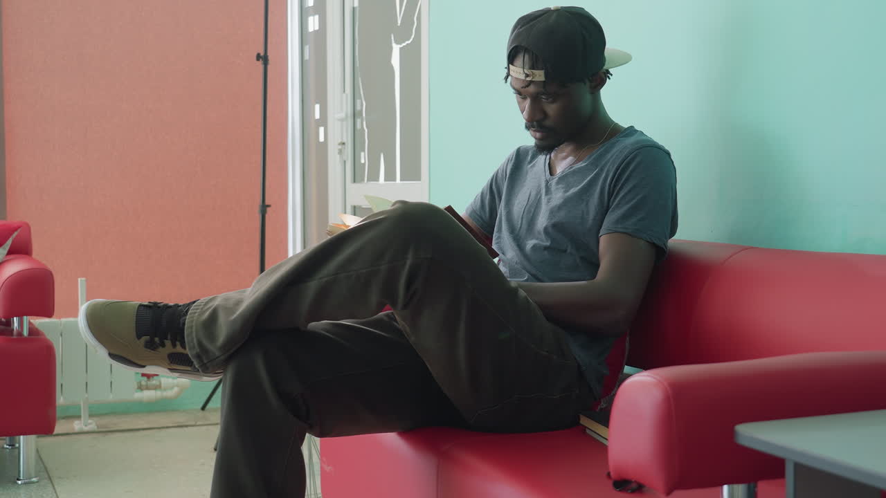Focused young man in casual outfit sitting on red couch, holding open book in hands and reading intently against plain teal wall, portraying calm indoor atmosphere