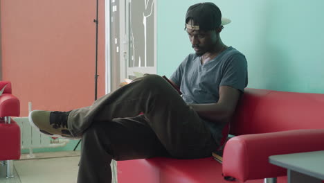 focused young man in casual outfit sitting on red couch, holding open book in hands and reading intently against plain teal wall, portraying calm indoor atmosphere