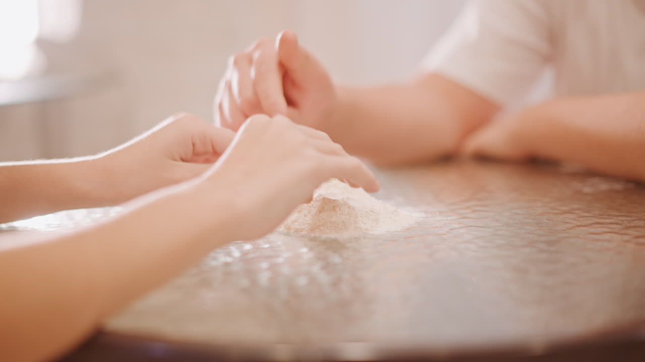 Mani bianche che spolverano farina sul tavolo, genitore e bambino che modellano un mucchio per l'impasto della torta di zucca in una cucina illuminata dal sole, gesti delicati di insegnamento, toni caldi, primo piano della texture della farina, movimento lento e attento