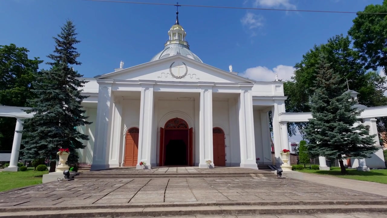 Edificio histórico de iglesia católica se alza bajo un cielo azul en Daugavpils en un soleado día de verano. Un plano general en contrapicado captura la gran fachada clásica y las columnas blancas con un ambiente sereno.