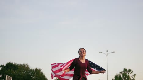 Junges-Glückliches-Amerikanisches-Mädchen,-Das-Läuft,-Während-Es-Die-Amerikanische-Flagge-Hält-Und-In-Die-Kamera-Schaut-1