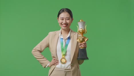 금메달과 트로피를 입은 슈트를 입은 아시아 사업가 여성이 한 팔로 카메라를 향해 미소 짓고 있으며, 아보는 스튜디오의 녹색 스크린 배경에서 첫 번째 우승자로 포즈를 취하고 있습니다.