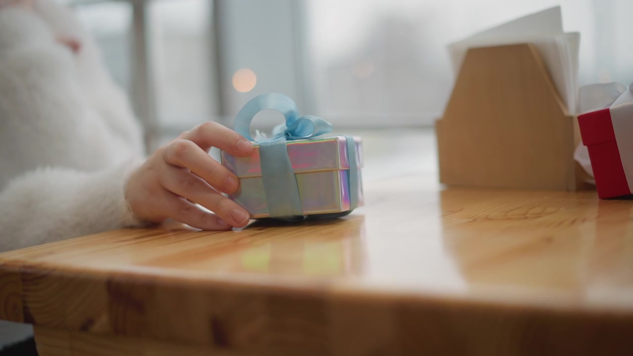 Vista em close-up de baixo ângulo de mãos colocando delicadamente uma pequena caixa de presente azul pastel com fita branca sobre uma mesa de madeira polida ao lado de uma caixa de papelão aberta; a ação visível mostra o contato cuidadoso das pontas dos dedos