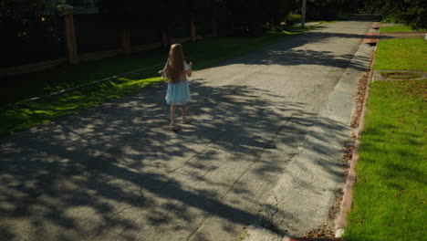 little girl in gown walking along cracked suburban street under leafy tree shade, surrounded by green grass, cement sidewalk, residential fences, and soft morning sunlight casting detailed shadows