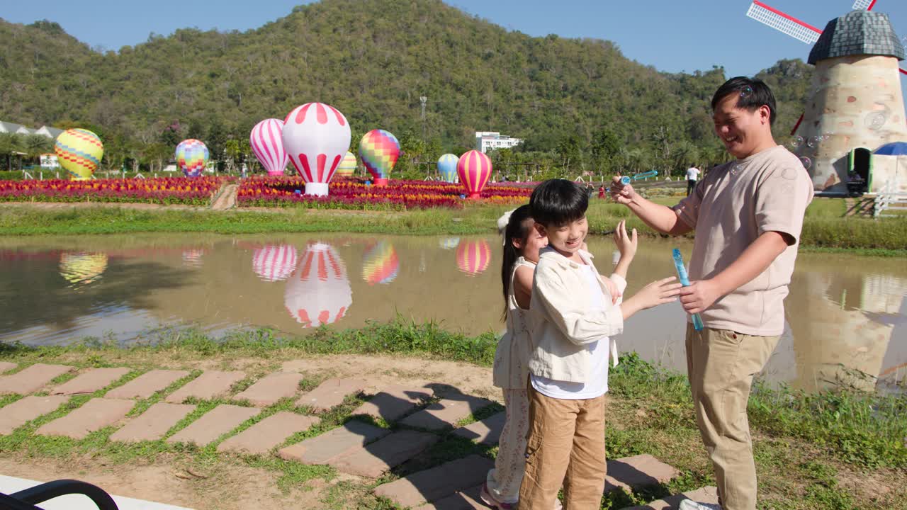 พ่อและลูกสองคนกำลังเป่าฟองสบู่อยู่นอกบ้านใกล้บ่อน้ำ โดยมีบอลลูนลมร้อนอยู่