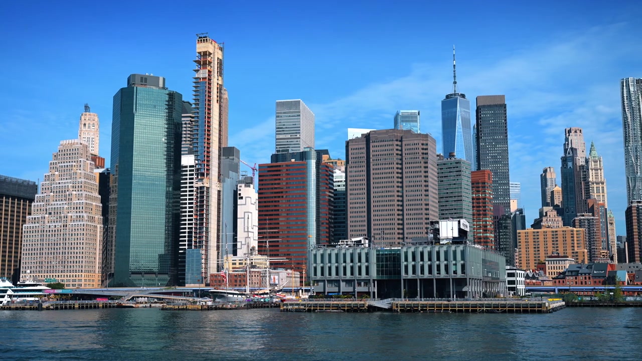 Horizonte do Distrito Financeiro visto do East River. Torres de escritórios iluminadas pelo sol se erguem atrás de barcos e um amplo píer à beira-mar sob um céu azul