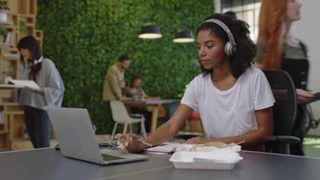 Joven-Mujer-De-Negocios-Afroamericana-Escribiendo-Notas-Disfrutando-De-Estudiar-Escuchando-Música-Estudiante-Negra-Usando-Una-Computadora-Portátil-Haciendo-Investigaciones-En-Línea-En-Un-Ocupado-Lugar-De-Trabajo-De-Oficina