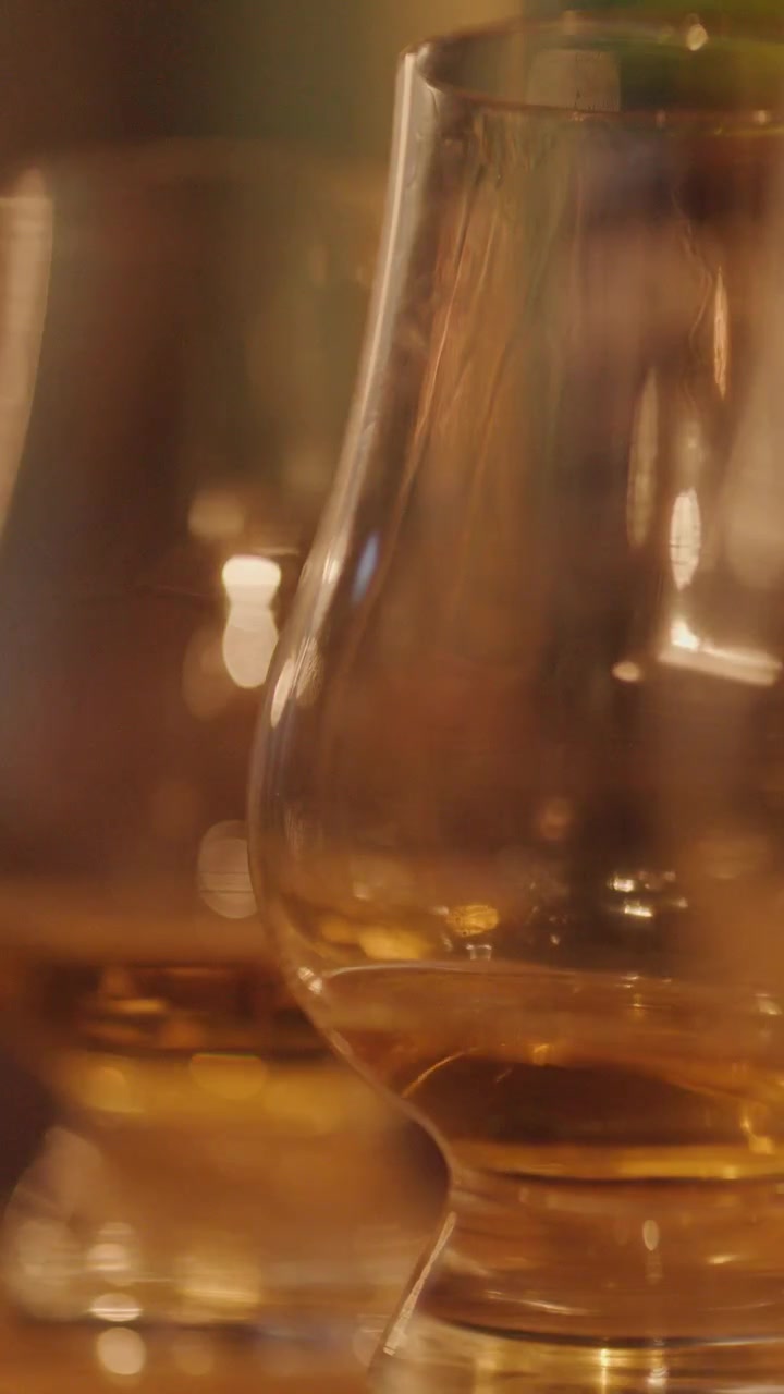 Una mano solleva un bicchiere di whisky da un bancone di legno, con una calda illuminazione ambientale che si riflette sul bicchiere e bottiglie leggermente sfocate sullo sfondo