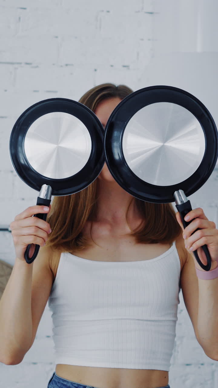 Young female with frypan in the kitchen. Attractive housewife holding two new nonstick pans and closing her face by them. Happy woman in the modern kitchen. Vertical video
