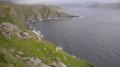 Wide-Shot-of-Puffins-Flying-Along-the-Cliffs-of-Runde-Norway-with-Mountains-in-the-Background,-Slow-Motion