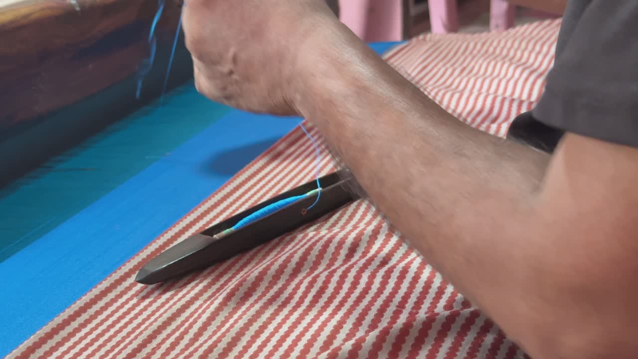 A closeup of a handloom worker's hand tying a knot to a thread using a specific knotting technique, to join two pieces of yarn or thread together on a loom