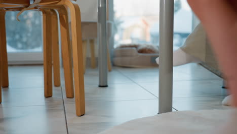partial view of woman's crossed leg with two other persons walking past and sitting down on a chair, all wearing white socks, the cozy indoor setting with white-tiled floor and minimal furniture