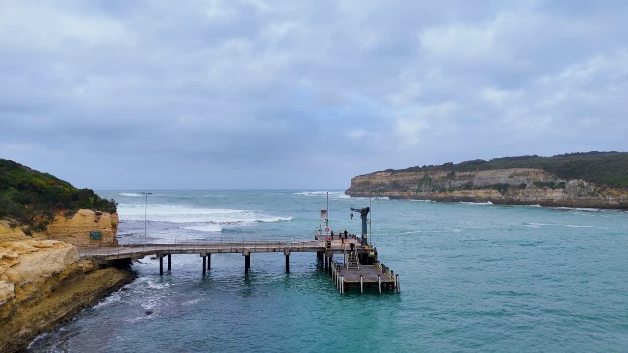 Drone footage captures Port Campbell's pier and surrounding bay under cloudy skies, highlighting coastal erosion and ocean waves