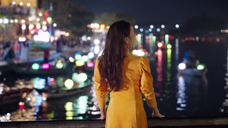 La-Mujer-De-Amarillo-Ao-Dai-Admira-La-Vibrante-Vida-Nocturna-Junto-Al-Río-En-Hoi-An,-Vietnam,-Con-Coloridas-Linternas-Reflejadas-En-El-Agua