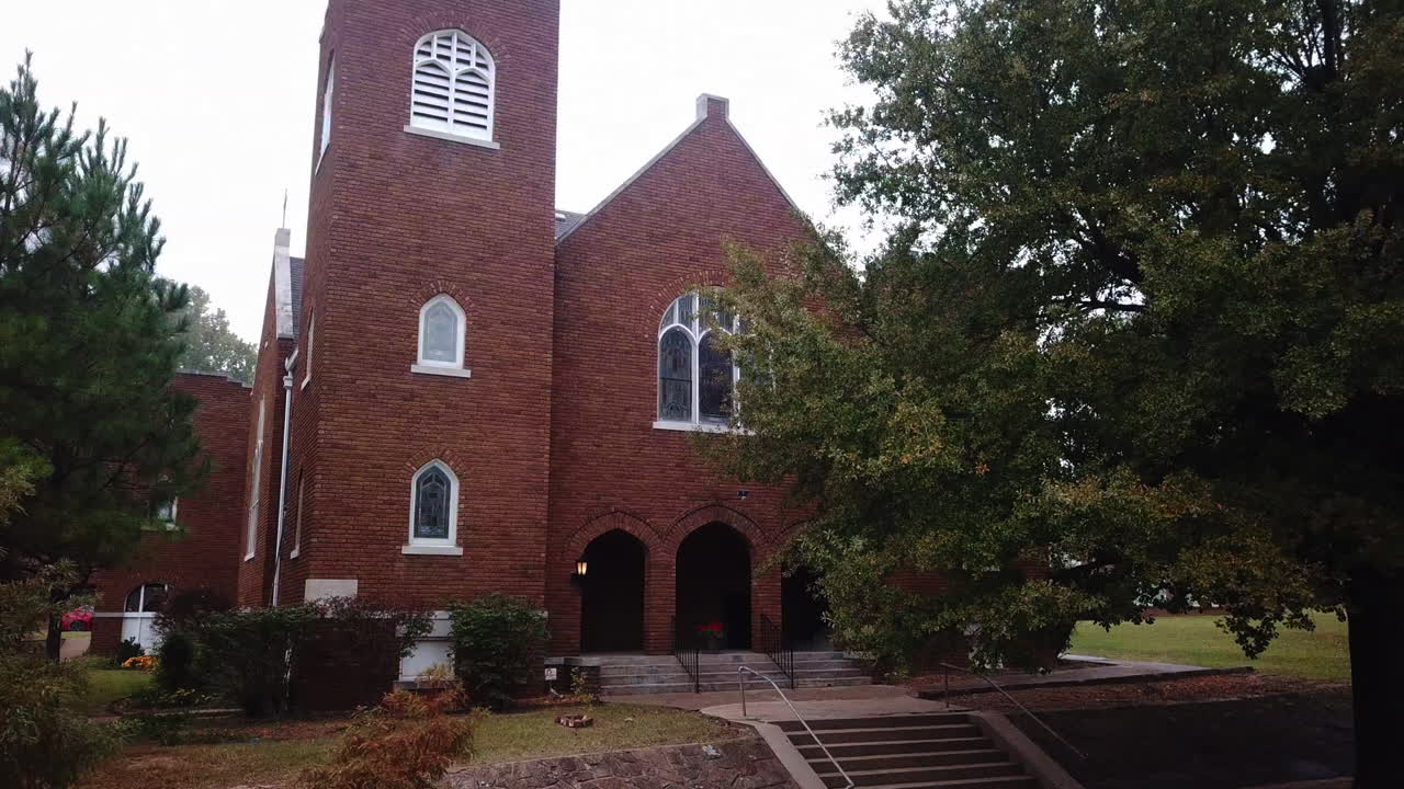 Drone footage from right to left of an old town with red brick walled church surrounded by giant trees