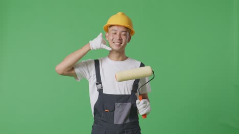 asian man painter wearing safety helmet smiling and making call me gesture to camera while standing in the green screen background studio