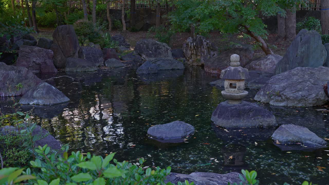 A serene, close-up view of a Japanese rock garden pond, featuring a stone lantern and mossy rocks reflected in the dark water