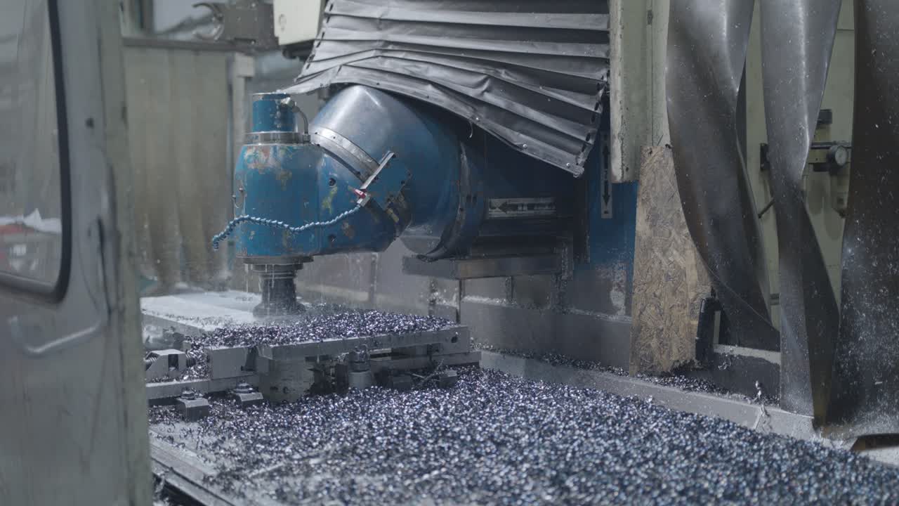 카메라는 금속 조각으로 가득 찬 컨베이어 벨트의 클로즈업에서 부드럽게 위로 기울어 고정밀 밀링 기계가 철판을 전문적으로 조각하는 것을 보여줍니다.