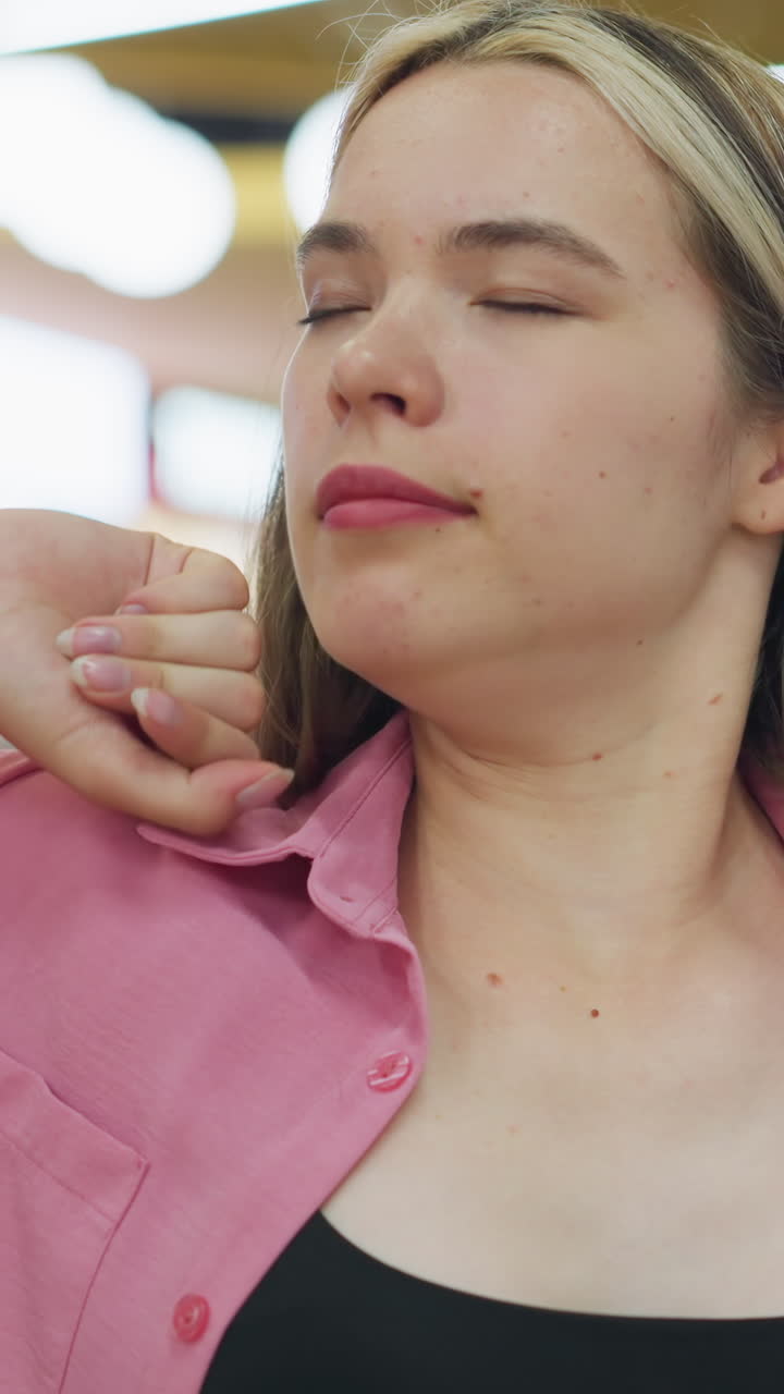 dama de vestido rosa que parece aburrida toma una respiración profunda y estira su cuello inclinando su cabeza de un lado a otro mientras coloca sus manos en su cuello, en el fondo, un hombre de negro pasa