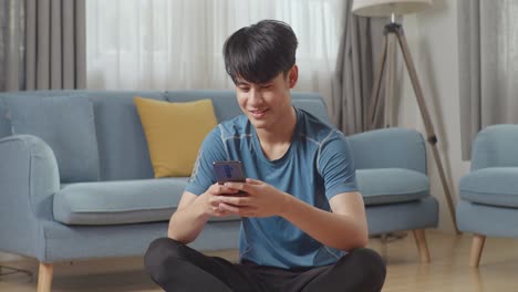 스포츠 옷을 입은 행복하고 운동적인 아시아인 남자가 집에서 아침 운동을 한 후 휴대전화를 사용하고 있습니다.