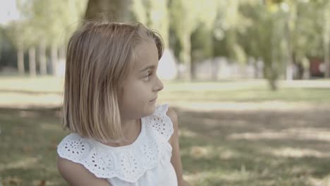 cute blonde little girl looking away and pointing at something while sitting on grass in the park