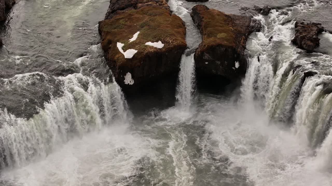 Close aerial view of Goðafoss waterfall in northern Iceland, showing powerful water streams splitting around mossy rocks, creating dramatic mist and turbulence.