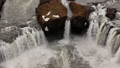 close aerial view of goðafoss waterfall in northern iceland, showing powerful water streams splitting around mossy rocks, creating dramatic mist and turbulence.