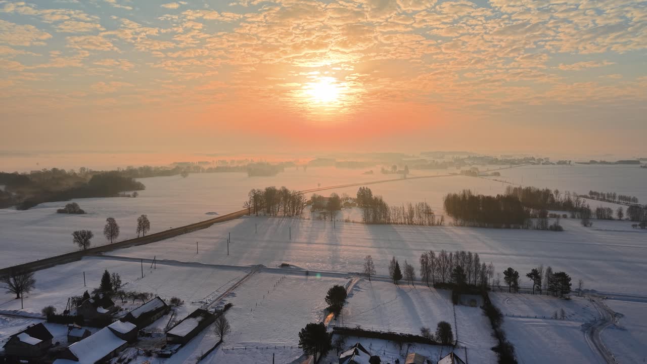Flyfoto fanger et fredfullt snødekt landskap ved soloppgang, med gyllent lys som kaster lange skygger over åkre og hustak, og viser en fantastisk vinterstillhet.