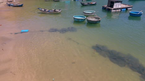 Coracle,-basket-boats-and-fishing-boats-aerial-fly-over-sandy-shore-and-aqua-green-blue-sea-waters-of-fishing-village,-Vietnam