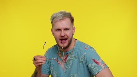 Seductive-cheerful-stylish-man-in-blue-t-shirt-wearing-sunglasses,-charming-smile-on-yellow-wall