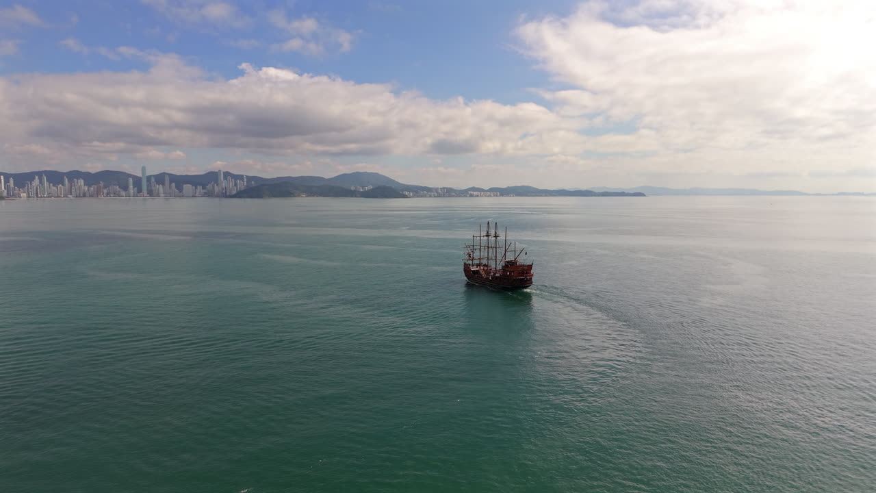 Lone pirate ship floating on calm ocean under dramatic sky with distant Balneário Camboriú city, Santa Catarina, Brazil