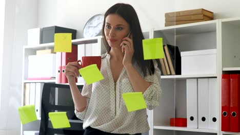 businesswoman in white blouse considering sticky notes on glass and talking on phone